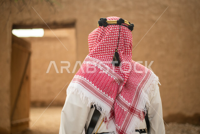 تصوير من الخلف لرجل عربي سعودي خليجي يرتدي الزي السعودي الشعبي التقليدي, الزي التراثي للمناسبات الوطنيه, التجول في إحدى المباني الطينيه الشعبيه, بيوت تراثيه قديمه, شعبيات, أحياء أثريه قديمه, جوله بين الأحياء الشعبيه
