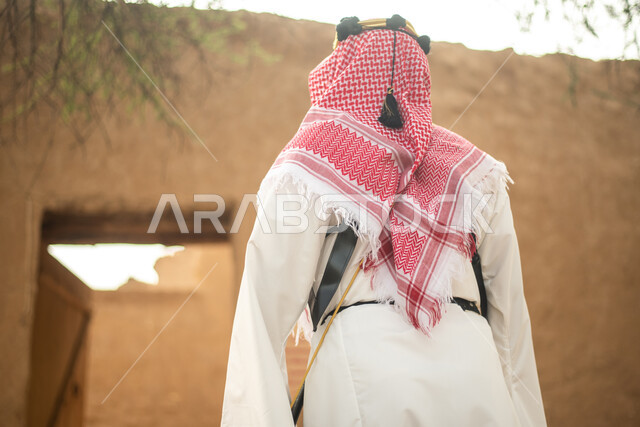 تصوير من الخلف لرجل عربي سعودي خليجي يرتدي الزي السعودي الشعبي التقليدي, الزي التراثي للمناسبات الوطنيه, التجول في إحدى المباني الطينيه الشعبيه, بيوت تراثيه قديمه, شعبيات, أحياء أثريه قديمه, جوله بين الأحياء الشعبيه