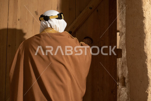 تصوير من الخلف لرجل مسن عربي سعودي خليجي يرتدي الزي السعودي الشعبي التقليدي, الزي التراثي للمناسبات الوطنيه, التجول في إحدى المباني الطينيه الشعبيه, بيوت تراثيه قديمه, شعبيات, أحياء أثريه قديمه, جوله بين الأحياء الشعبيه