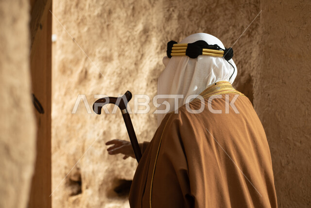 تصوير من الخلف لرجل مسن عربي سعودي خليجي يرتدي الزي السعودي الشعبي التقليدي, الزي التراثي للمناسبات الوطنيه, التجول في إحدى المباني الطينيه الشعبيه, بيوت تراثيه قديمه, شعبيات, أحياء أثريه قديمه, جوله بين الأحياء الشعبيه