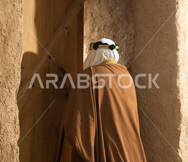 تصوير من الخلف لرجل مسن عربي سعودي خليجي يرتدي الزي السعودي الشعبي التقليدي, الزي التراثي للمناسبات الوطنيه, التجول في إحدى المباني الطينيه الشعبيه, بيوت تراثيه قديمه, شعبيات, أحياء أثريه قديمه, جوله بين الأحياء الشعبيه