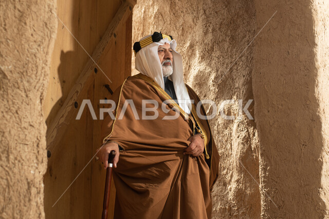 رجل مسن عربي سعودي خليجي يرتدي الزي السعودي الشعبي التقليدي, الزي التراثي للمناسبات الوطنيه, الوقوف في إحدى المباني الطينيه الشعبيه, بيوت تراثيه قديمه, شعبيات, يوم التأسيس السعودي, الإحتفالات الوطنيه في المنطقه الوسطى, مباني أثريه قديمه