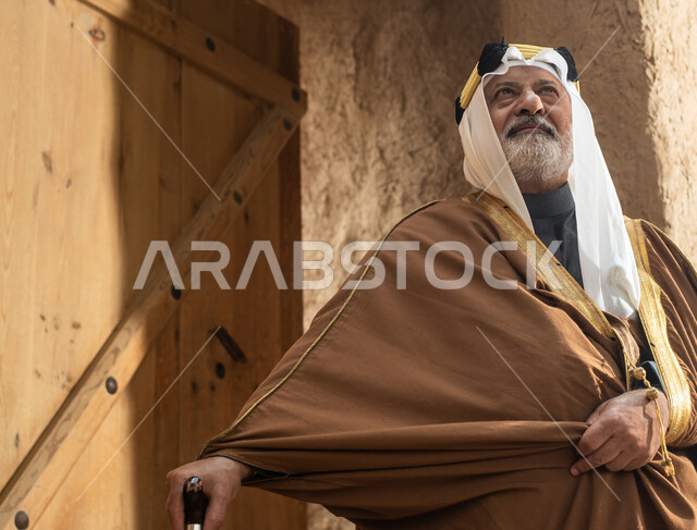 رجل مسن عربي سعودي خليجي يرتدي الزي السعودي الشعبي التقليدي, الزي التراثي للمناسبات الوطنيه, الوقوف في إحدى المباني الطينيه الشعبيه, بيوت تراثيه قديمه, شعبيات, يوم التأسيس السعودي, الإحتفالات الوطنيه في المنطقه الوسطى, مباني أثريه قديمه