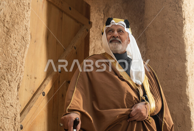 رجل مسن عربي سعودي خليجي يرتدي الزي السعودي الشعبي التقليدي, الزي التراثي للمناسبات الوطنيه, الوقوف في إحدى المباني الطينيه الشعبيه, بيوت تراثيه قديمه, شعبيات, يوم التأسيس السعودي, الإحتفالات الوطنيه في المنطقه الوسطى, مباني أثريه قديمه