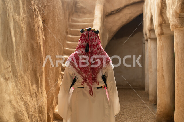 تصوير من الخلف لرجل عربي سعودي خليجي يرتدي الزي السعودي الشعبي التقليدي, الزي التراثي للمناسبات الوطنيه, التجول في إحدى المباني الطينيه الشعبيه, بيوت تراثيه قديمه, شعبيات, أحياء أثريه قديمه, جوله بين الأحياء الشعبيه