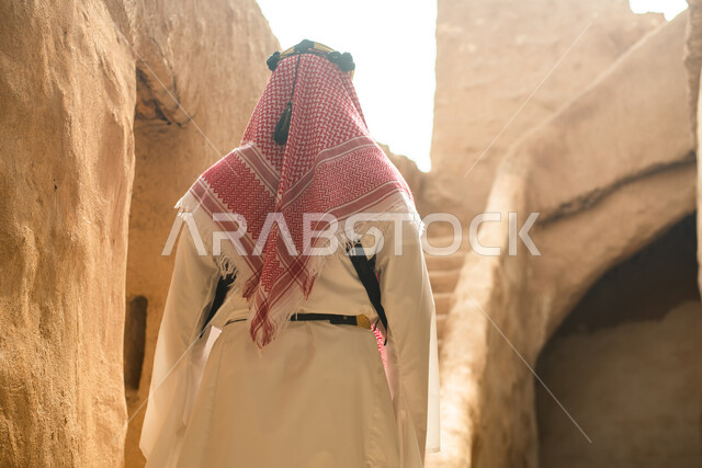 Photography from the back of a Saudi Arabian Gulf man wearing the traditional Saudi folk costume, the traditional costume for national occasions, touring in one of the popular mud buildings, old heritage houses, folklore, ancient ancient neighborhoods, a 