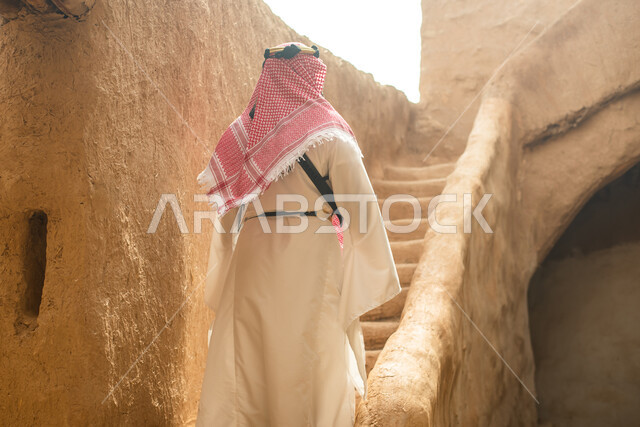 تصوير من الخلف لرجل عربي سعودي خليجي يرتدي الزي السعودي الشعبي التقليدي, الزي التراثي للمناسبات الوطنيه, التجول في إحدى المباني الطينيه الشعبيه, بيوت تراثيه قديمه, شعبيات, أحياء أثريه قديمه, جوله بين الأحياء الشعبيه