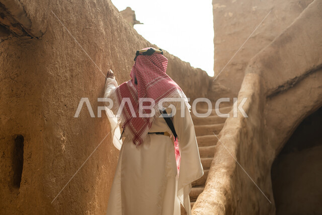تصوير من الخلف لرجل عربي سعودي خليجي يرتدي الزي السعودي الشعبي التقليدي, الزي التراثي للمناسبات الوطنيه, التجول في إحدى المباني الطينيه الشعبيه, بيوت تراثيه قديمه, شعبيات, أحياء أثريه قديمه, جوله بين الأحياء الشعبيه