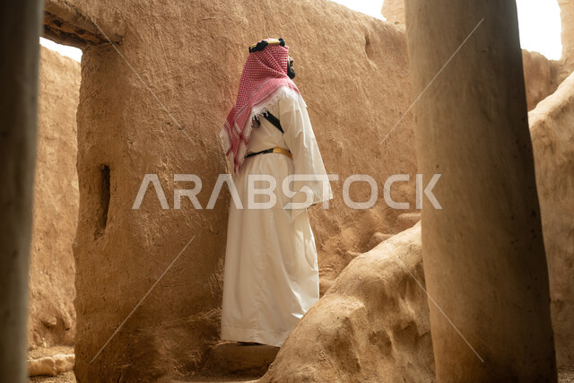تصوير من الخلف لرجل عربي سعودي خليجي يرتدي الزي السعودي الشعبي التقليدي, الزي التراثي للمناسبات الوطنيه, التجول في إحدى المباني الطينيه الشعبيه, بيوت تراثيه قديمه, شعبيات, أحياء أثريه قديمه, جوله بين الأحياء الشعبيه