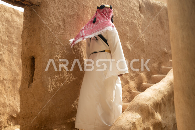Photography from the back of a Saudi Arabian Gulf man wearing the traditional Saudi folk costume, the traditional costume for national occasions, touring in one of the popular mud buildings, old heritage houses, folklore, ancient ancient neighborhoods, a 