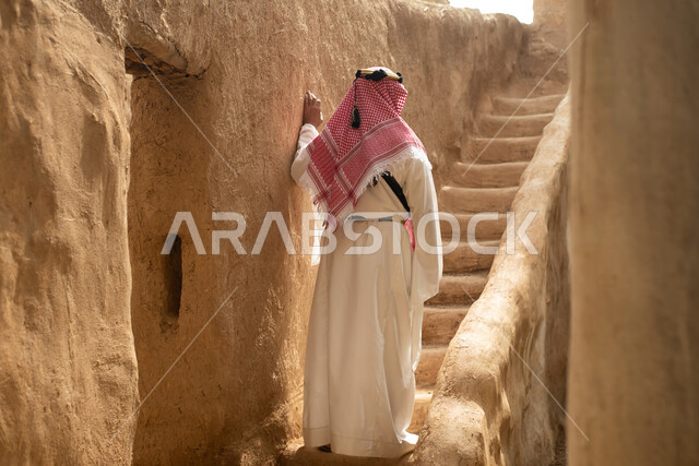 تصوير من الخلف لرجل عربي سعودي خليجي يرتدي الزي السعودي الشعبي التقليدي, الزي التراثي للمناسبات الوطنيه, التجول في إحدى المباني الطينيه الشعبيه, بيوت تراثيه قديمه, شعبيات, أحياء أثريه قديمه, جوله بين الأحياء الشعبيه