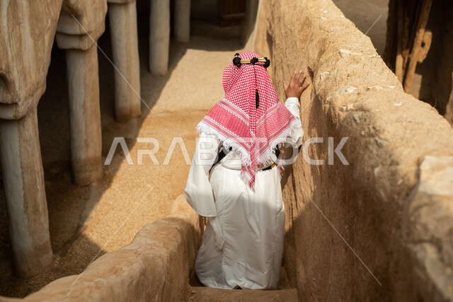 Photography from the back of a Saudi Arabian Gulf man wearing the traditional Saudi folk costume, the traditional costume for national occasions, touring in one of the popular mud buildings, old heritage houses, folklore, ancient ancient neighborhoods, a 