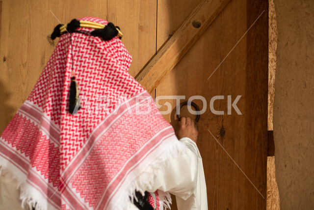 Photography from the back of a Saudi Arabian Gulf man wearing the traditional Saudi folk costume, the traditional costume for national occasions, touring in one of the popular mud buildings, old heritage houses, folklore, ancient ancient neighborhoods, a 