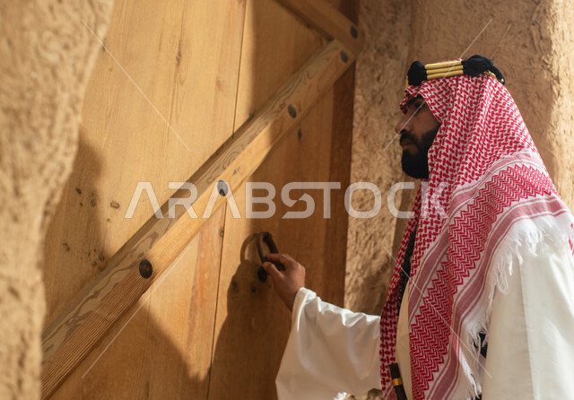 Photography from the back of a Saudi Arabian Gulf man wearing the traditional Saudi folk costume, the traditional costume for national occasions, touring in one of the popular mud buildings, old heritage houses, folklore, ancient ancient neighborhoods, a 