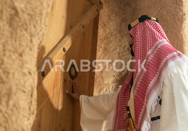 Photography from the back of a Saudi Arabian Gulf man wearing the traditional Saudi folk costume, the traditional costume for national occasions, touring in one of the popular mud buildings, old heritage houses, folklore, ancient ancient neighborhoods, a 