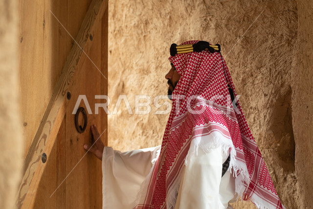 تصوير من الخلف لرجل عربي سعودي خليجي يرتدي الزي السعودي الشعبي التقليدي, الزي التراثي للمناسبات الوطنيه, التجول في إحدى المباني الطينيه الشعبيه, بيوت تراثيه قديمه, شعبيات, أحياء أثريه قديمه, جوله بين الأحياء الشعبيه