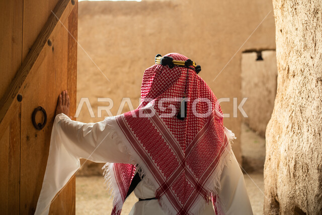 تصوير من الخلف لرجل عربي سعودي خليجي يرتدي الزي السعودي الشعبي التقليدي, الزي التراثي للمناسبات الوطنيه, التجول في إحدى المباني الطينيه الشعبيه, بيوت تراثيه قديمه, شعبيات, أحياء أثريه قديمه, جوله بين الأحياء الشعبيه