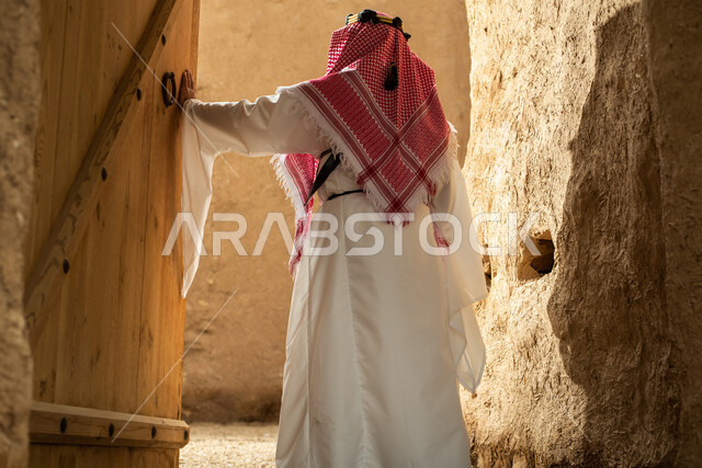 تصوير من الخلف لرجل عربي سعودي خليجي يرتدي الزي السعودي الشعبي التقليدي, الزي التراثي للمناسبات الوطنيه, التجول في إحدى المباني الطينيه الشعبيه, بيوت تراثيه قديمه, شعبيات, أحياء أثريه قديمه, جوله بين الأحياء الشعبيه