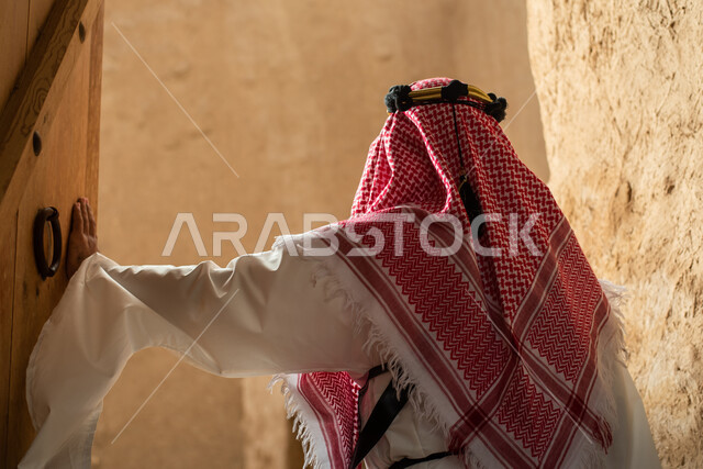 Photography from the back of a Saudi Arabian Gulf man wearing the traditional Saudi folk costume, the traditional costume for national occasions, touring in one of the popular mud buildings, old heritage houses, folklore, ancient ancient neighborhoods, a 