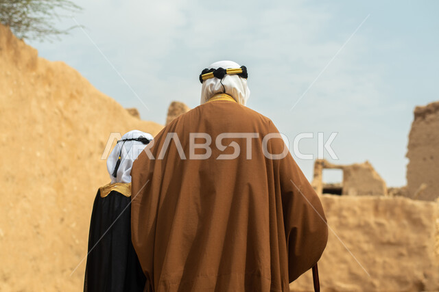 صوره من الخلف لرجل مسن وولد عربيان سعوديان خليجيان يرتديان الزي السعودي الشعبي التقليدي, الزي التراثي للمناسبات الوطنيه, التجول في إحدى المباني الطينيه الشعبيه, بيوت تراثيه قديمه, شعبيات, أحياء أثريه قديمه, جوله بين الأحياء الشعبيه
