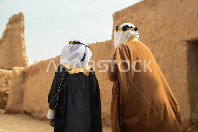 صوره من الخلف لرجل مسن وولد عربيان سعوديان خليجيان يرتديان الزي السعودي الشعبي التقليدي, الزي التراثي للمناسبات الوطنيه, التجول في إحدى المباني الطينيه الشعبيه, بيوت تراثيه قديمه, شعبيات, أحياء أثريه قديمه, جوله بين الأحياء الشعبيه
