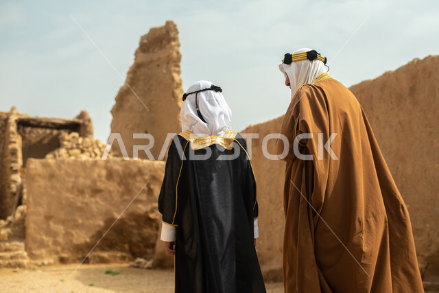A picture from the back of an elderly man and a boy, two Saudi Arabs from the Gulf, dressed in the traditional Saudi folk dress, the traditional dress for national occasions, touring in one of the popular mud buildings, old heritage houses, folklore, anci