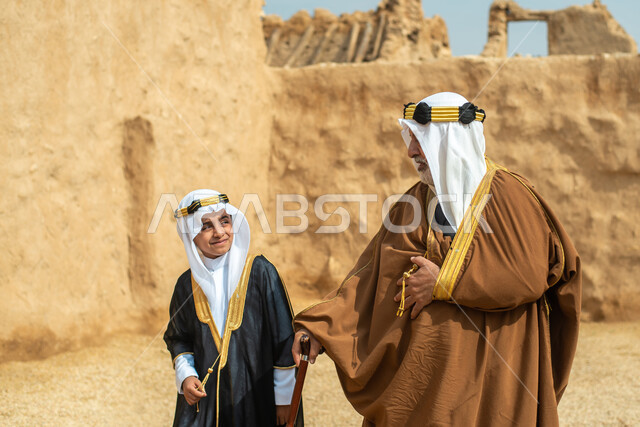 رجل مسن وولد عربيان سعوديان خليجيان يرتديان الزي السعودي الشعبي التقليدي, الزي التراثي للمناسبات الوطنيه, التجول في إحدى المباني الطينيه الشعبيه, بيوت تراثيه قديمه, شعبيات, أحياء أثريه قديمه, جوله بين الأحياء الشعبيه