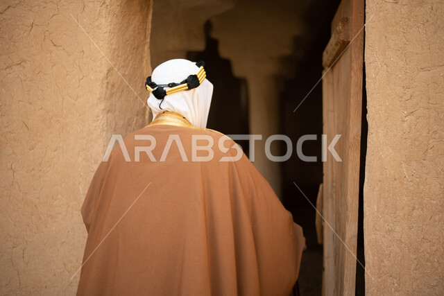 تصوير من الخلف لرجل مسن عربي سعودي خليجي يرتدي الزي السعودي الشعبي التقليدي, الزي التراثي للمناسبات الوطنيه, التجول في إحدى المباني الطينيه الشعبيه, بيوت تراثيه قديمه, شعبيات, أحياء أثريه قديمه, جوله بين الأحياء الشعبيه