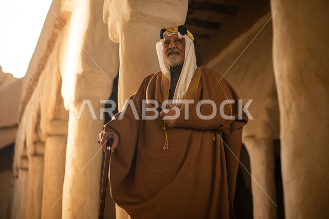 رجل مسن عربي سعودي خليجي يرتدي الزي السعودي الشعبي التقليدي, الزي التراثي للمناسبات الوطنيه, التجول في إحدى المباني الطينيه الشعبيه, بيوت تراثيه قديمه, شعبيات, أحياء أثريه قديمه, جوله بين الأحياء الشعبيه