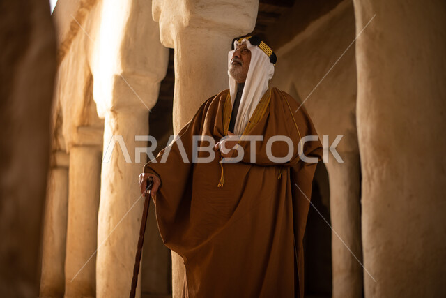 رجل مسن عربي سعودي خليجي يرتدي الزي السعودي الشعبي التقليدي, الزي التراثي للمناسبات الوطنيه, التجول في إحدى المباني الطينيه الشعبيه, بيوت تراثيه قديمه, شعبيات, أحياء أثريه قديمه, جوله بين الأحياء الشعبيه