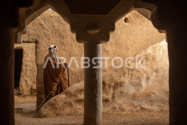 رجل مسن عربي سعودي خليجي يرتدي الزي السعودي الشعبي التقليدي, الزي التراثي للمناسبات الوطنيه, التجول في إحدى المباني الطينيه الشعبيه, بيوت تراثيه قديمه, شعبيات, أحياء أثريه قديمه, جوله بين الأحياء الشعبيه