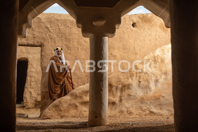 رجل مسن عربي سعودي خليجي يرتدي الزي السعودي الشعبي التقليدي, الزي التراثي للمناسبات الوطنيه, التجول في إحدى المباني الطينيه الشعبيه, بيوت تراثيه قديمه, شعبيات, أحياء أثريه قديمه, جوله بين الأحياء الشعبيه
