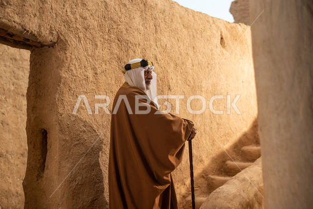 تصوير من الخلف لرجل مسن عربي سعودي خليجي يرتدي الزي السعودي الشعبي التقليدي, الزي التراثي للمناسبات الوطنيه, التجول في إحدى المباني الطينيه الشعبيه, بيوت تراثيه قديمه, شعبيات, أحياء أثريه قديمه, جوله بين الأحياء الشعبيه
