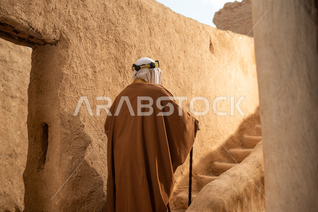 تصوير من الخلف لرجل مسن عربي سعودي خليجي يرتدي الزي السعودي الشعبي التقليدي, الزي التراثي للمناسبات الوطنيه, التجول في إحدى المباني الطينيه الشعبيه, بيوت تراثيه قديمه, شعبيات, أحياء أثريه قديمه, جوله بين الأحياء الشعبيه