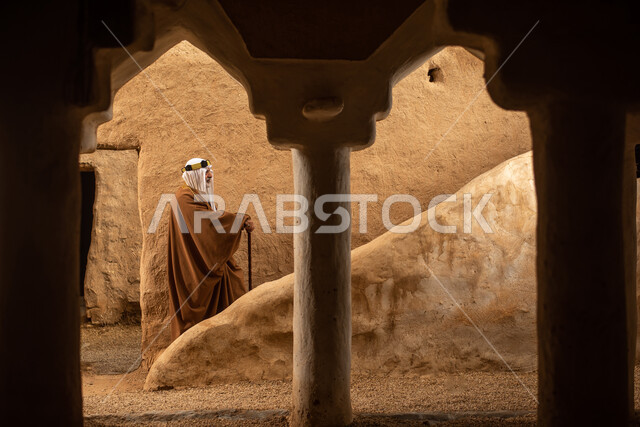 تصوير من الخلف لرجل مسن عربي سعودي خليجي يرتدي الزي السعودي الشعبي التقليدي, الزي التراثي للمناسبات الوطنيه, التجول في إحدى المباني الطينيه الشعبيه, بيوت تراثيه قديمه, شعبيات, أحياء أثريه قديمه, جوله بين الأحياء الشعبيه