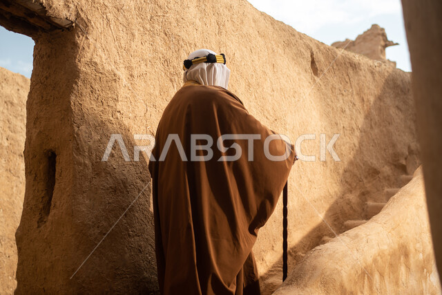 تصوير من الخلف لرجل مسن عربي سعودي خليجي يرتدي الزي السعودي الشعبي التقليدي, الزي التراثي للمناسبات الوطنيه, التجول في إحدى المباني الطينيه الشعبيه, بيوت تراثيه قديمه, شعبيات, أحياء أثريه قديمه, جوله بين الأحياء الشعبيه