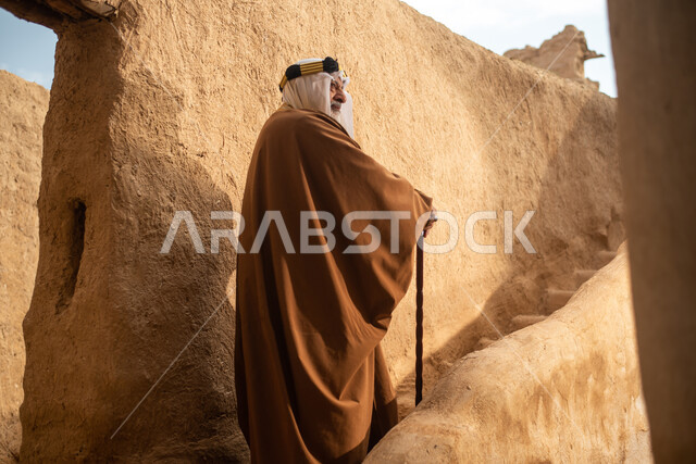تصوير من الخلف لرجل مسن عربي سعودي خليجي يرتدي الزي السعودي الشعبي التقليدي, الزي التراثي للمناسبات الوطنيه, التجول في إحدى المباني الطينيه الشعبيه, بيوت تراثيه قديمه, شعبيات, أحياء أثريه قديمه, جوله بين الأحياء الشعبيه