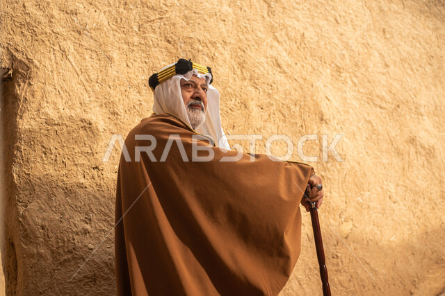 تصوير من الخلف لرجل مسن عربي سعودي خليجي يرتدي الزي السعودي الشعبي التقليدي, الزي التراثي للمناسبات الوطنيه, التجول في إحدى المباني الطينيه الشعبيه, بيوت تراثيه قديمه, شعبيات, أحياء أثريه قديمه, جوله بين الأحياء الشعبيه