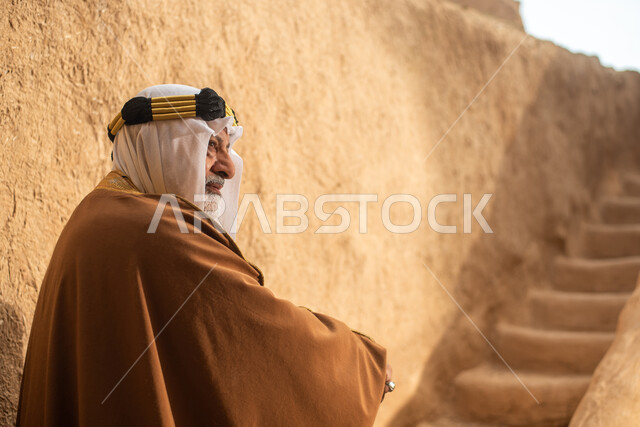 تصوير من الخلف لرجل مسن عربي سعودي خليجي يرتدي الزي السعودي الشعبي التقليدي, الزي التراثي للمناسبات الوطنيه, التجول في إحدى المباني الطينيه الشعبيه, بيوت تراثيه قديمه, شعبيات, أحياء أثريه قديمه, جوله بين الأحياء الشعبيه