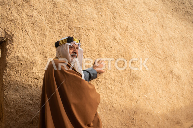 رجل مسن عربي سعودي خليجي يرتدي الزي السعودي الشعبي التقليدي, الزي التراثي للمناسبات الوطنيه, التجول في إحدى المباني الطينيه الشعبيه, بيوت تراثيه قديمه, شعبيات, أحياء أثريه قديمه, جوله بين الأحياء الشعبيه