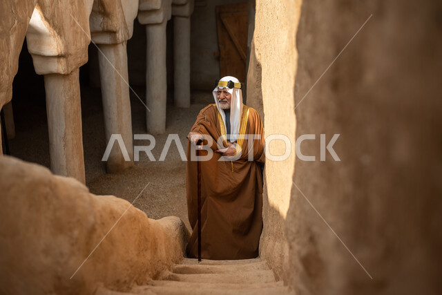 رجل مسن عربي سعودي خليجي يرتدي الزي السعودي الشعبي التقليدي, الزي التراثي للمناسبات الوطنيه, التجول في إحدى المباني الطينيه الشعبيه, بيوت تراثيه قديمه, شعبيات, أحياء أثريه قديمه, جوله بين الأحياء الشعبيه