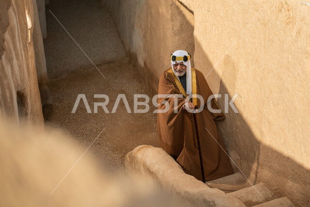 رجل مسن عربي سعودي خليجي يرتدي الزي السعودي الشعبي التقليدي, الزي التراثي للمناسبات الوطنيه, التجول في إحدى المباني الطينيه الشعبيه, بيوت تراثيه قديمه, شعبيات, أحياء أثريه قديمه, جوله بين الأحياء الشعبيه