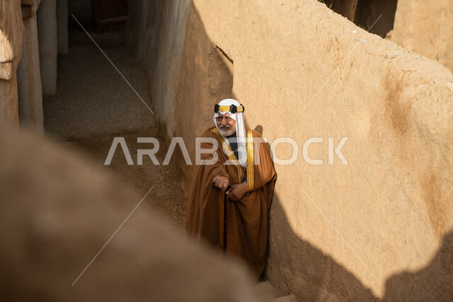 رجل مسن عربي سعودي خليجي يرتدي الزي السعودي الشعبي التقليدي, الزي التراثي للمناسبات الوطنيه, التجول في إحدى المباني الطينيه الشعبيه, بيوت تراثيه قديمه, شعبيات, أحياء أثريه قديمه, جوله بين الأحياء الشعبيه