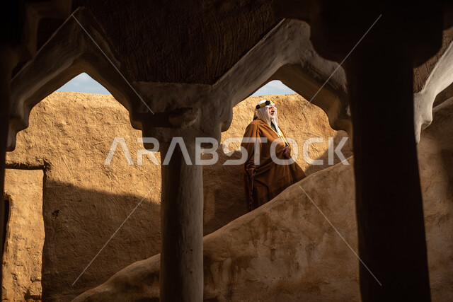 رجل مسن عربي سعودي خليجي يرتدي الزي السعودي الشعبي التقليدي, الزي التراثي للمناسبات الوطنيه, التجول في إحدى المباني الطينيه الشعبيه, بيوت تراثيه قديمه, شعبيات, أحياء أثريه قديمه, جوله بين الأحياء الشعبيه