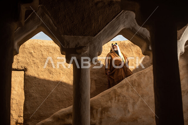 رجل مسن عربي سعودي خليجي يرتدي الزي السعودي الشعبي التقليدي, الزي التراثي للمناسبات الوطنيه, التجول في إحدى المباني الطينيه الشعبيه, بيوت تراثيه قديمه, شعبيات, أحياء أثريه قديمه, جوله بين الأحياء الشعبيه