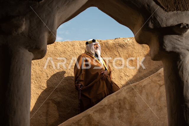 رجل مسن عربي سعودي خليجي يرتدي الزي السعودي الشعبي التقليدي, الزي التراثي للمناسبات الوطنيه, التجول في إحدى المباني الطينيه الشعبيه, بيوت تراثيه قديمه, شعبيات, أحياء أثريه قديمه, جوله بين الأحياء الشعبيه