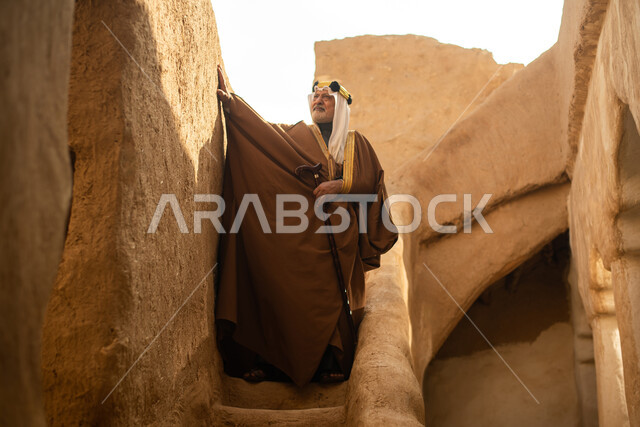 رجل مسن عربي سعودي خليجي يرتدي الزي السعودي الشعبي التقليدي, الزي التراثي للمناسبات الوطنيه, التجول في إحدى المباني الطينيه الشعبيه, بيوت تراثيه قديمه, شعبيات, أحياء أثريه قديمه, جوله بين الأحياء الشعبيه
