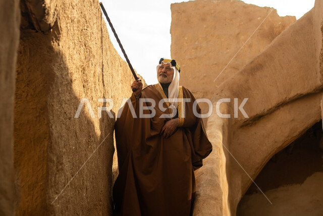 رجل مسن عربي سعودي خليجي يرتدي الزي السعودي الشعبي التقليدي, الزي التراثي للمناسبات الوطنيه, التجول في إحدى المباني الطينيه الشعبيه, بيوت تراثيه قديمه, شعبيات, أحياء أثريه قديمه, جوله بين الأحياء الشعبيه