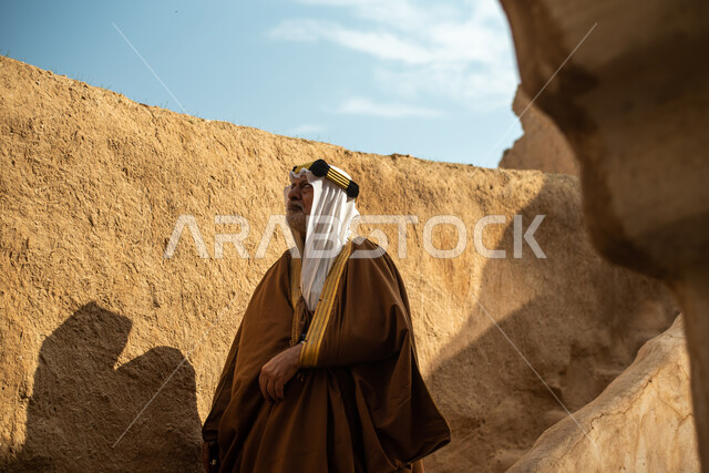 رجل مسن عربي سعودي خليجي يرتدي الزي السعودي الشعبي التقليدي, الزي التراثي للمناسبات الوطنيه, التجول في إحدى المباني الطينيه الشعبيه, بيوت تراثيه قديمه, شعبيات, أحياء أثريه قديمه, جوله بين الأحياء الشعبيه
