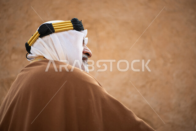 تصوير من الخلف لرجل مسن عربي سعودي خليجي يرتدي الزي السعودي الشعبي التقليدي, الزي التراثي للمناسبات الوطنيه, التجول في إحدى المباني الطينيه الشعبيه, بيوت تراثيه قديمه, شعبيات, أحياء أثريه قديمه, جوله بين الأحياء الشعبيه